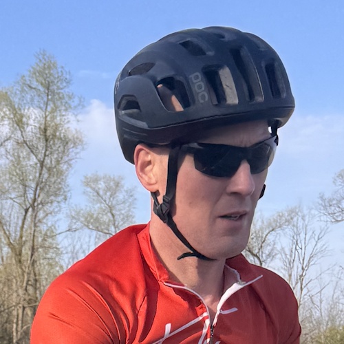 Angled profile of male road cyclist's headshot, complete with black helmet and sunglasses, wearing red cycling jersey.